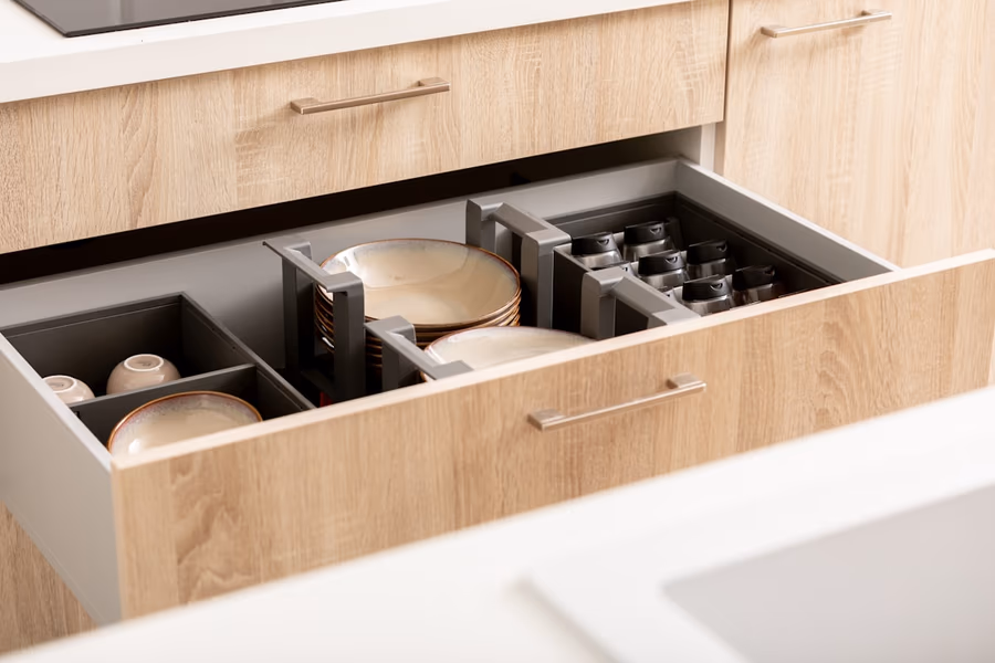 An open kitchen base cabinet drawer with dividers holding plates, bowls, and jars, beneath a wood-fronted upper drawer