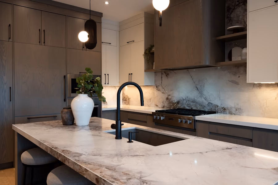 A kitchen island with a stone countertop and an undermount sink, with a wall-mounted range hood ducted above a gas range in the background