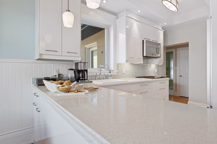 A peninsula with an engineered quartz countertop in a white kitchen, with an over-the-range microwave beyond and a window above the sink